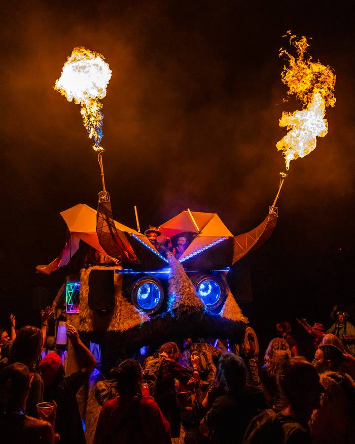 The Kuker at night with glowing blue eyes and flames from both horns above a dancing crowd