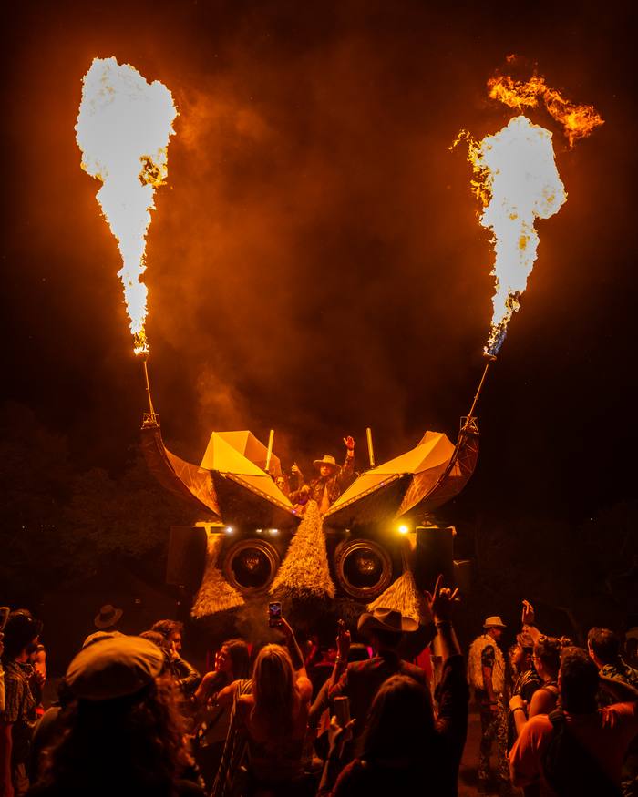 A DJ raising a hand in the Kuker's booth as tall flames rise from both horns over the crowd