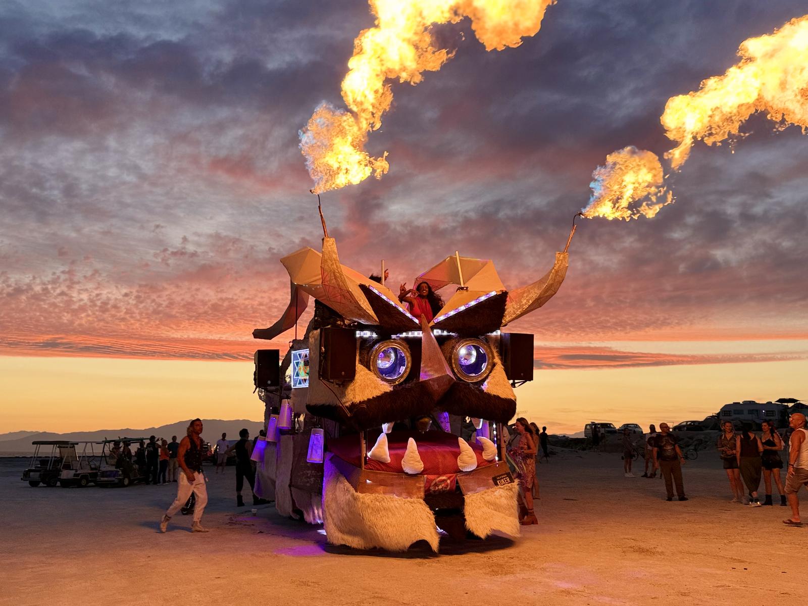 The Kuker art car at sunset in the desert, flames shooting from both horns, with DJs in the booth and people dancing around it